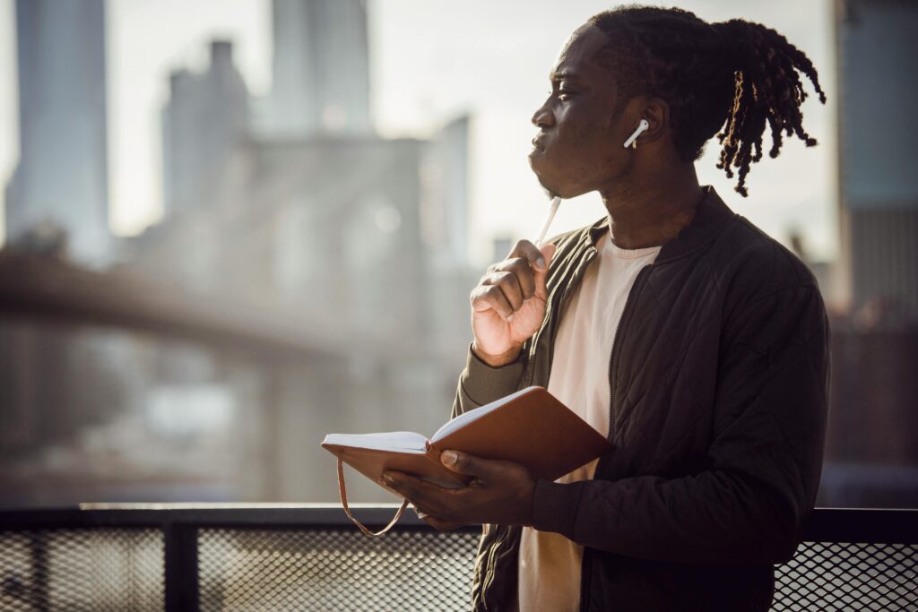 Young man with earbuds writing in a notepad outdoors, looking thoughtful, in a city setting.